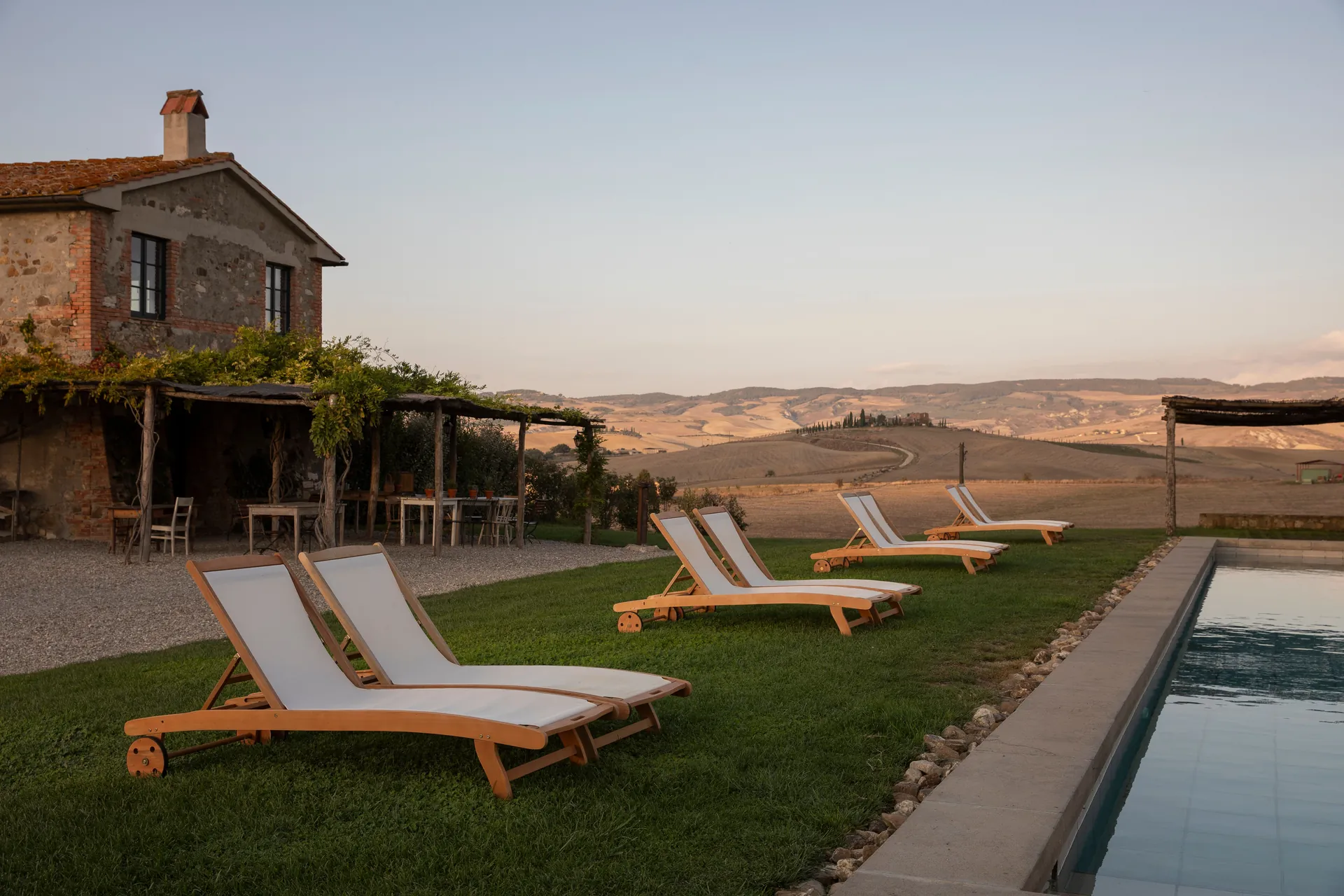 Il casale in pietra con il pergolato, le sdraio sul prato e la piscina affacciata sulle colline al tramonto